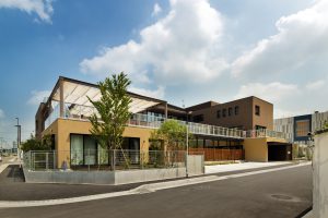 黄色と茶色の外壁を持つ介護施設の建物外観。現代的な設計で、エントランス周辺に植栽がある。