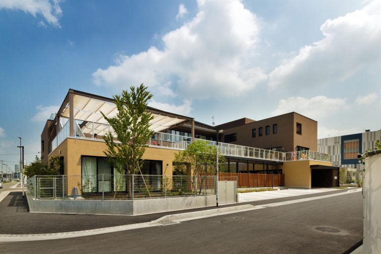 現代的な黄色と茶色の外壁を持つ介護施設の建物全景。広々とした敷地と緑化が見られます。